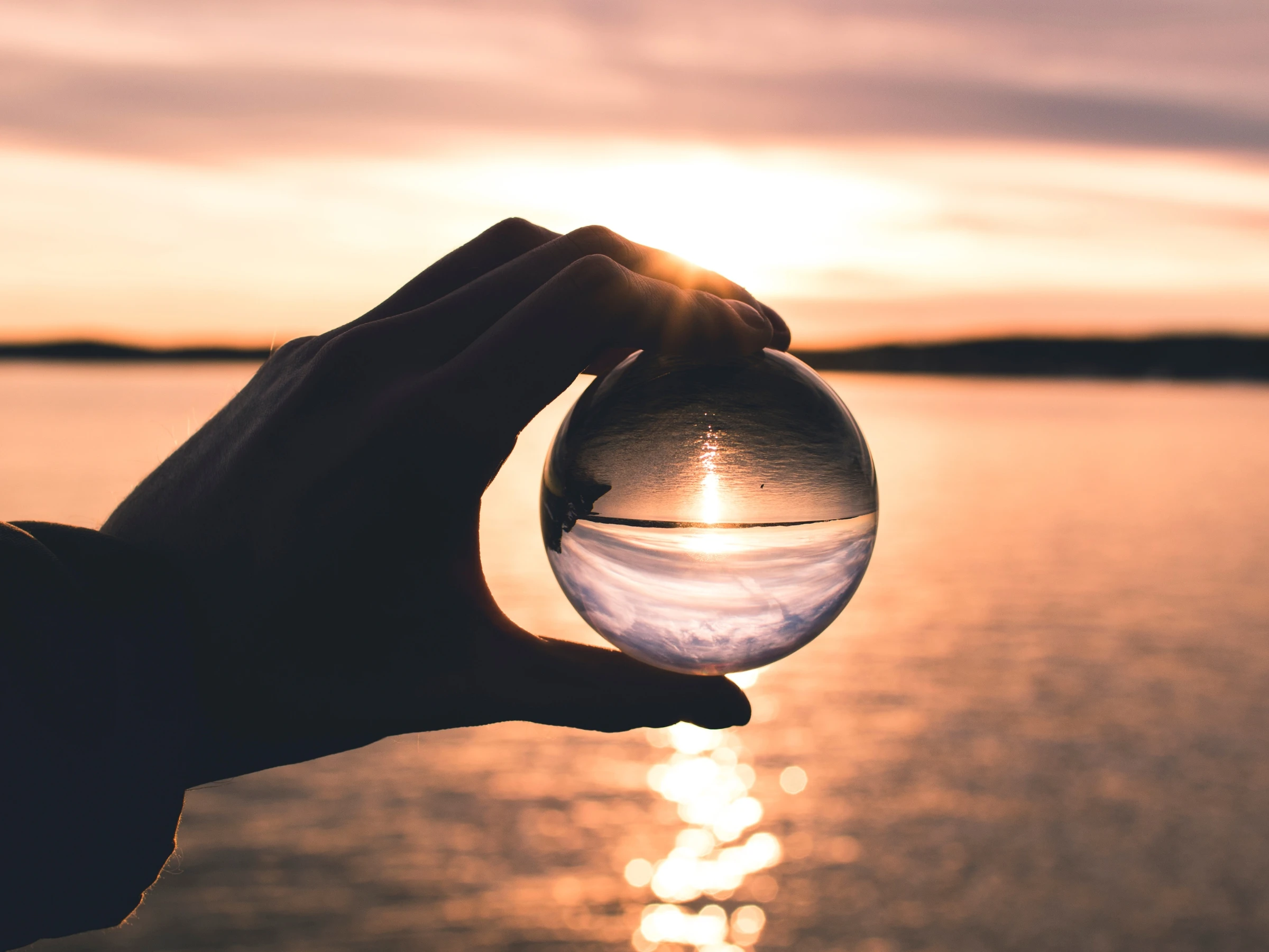 Hand holding a glass sphere with an inverted reflection of a sunset over a calm body of water.